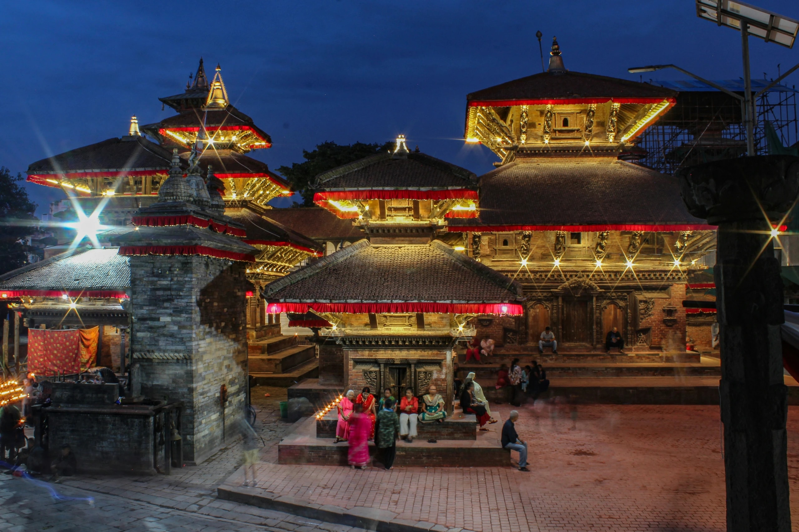 Traditional multi-tiered Nepalese Hindu temples illuminated at dusk with warm lights, as people gather in the courtyard.