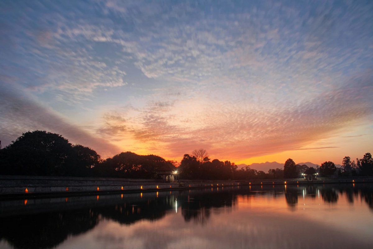 Vibrant orange sunset glowing over Siddha Pokhari pond in Bhaktapur, reflecting clouds and tree silhouettes on calm water.
